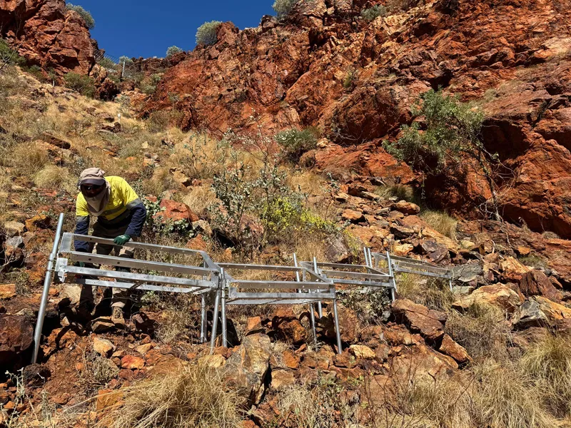 Field worker carrying a metal scaffold structure across red-rock terrain for a remote installation.